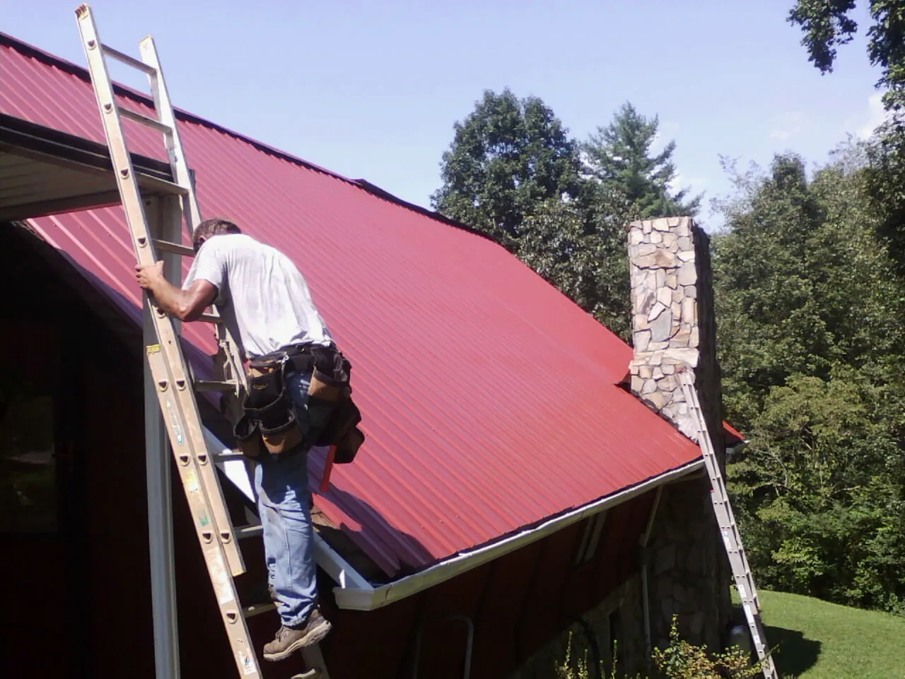 Roofer on a ladder beside a red metal roof and stone chimney — Soffit & Fascia Repair by Roofing Brothers in Maiden